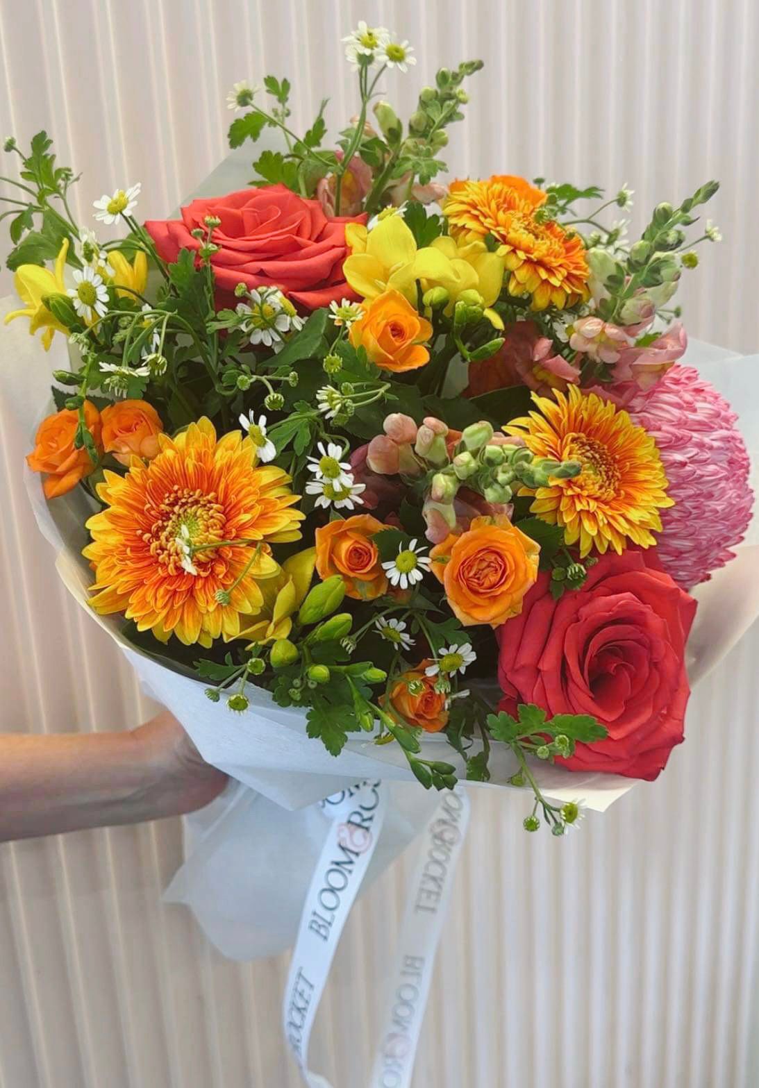 A bouquet of bright flowers including red roses and orange gerberas, wrapped in white paper with a ribbon. Bloom and Rocket Florist, Taringa. Brisbane Flower Delivery.