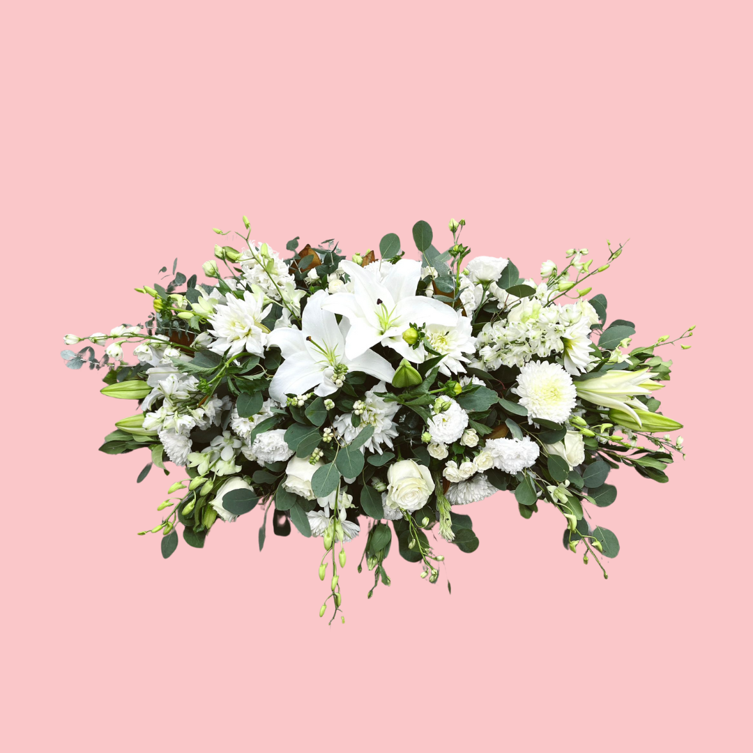 A floral casket funeral spray with a variety of flowers including white lilies and green foliage, arranged on a pink background. Brisbane Florist. Bloom and Rocket Taringa.
