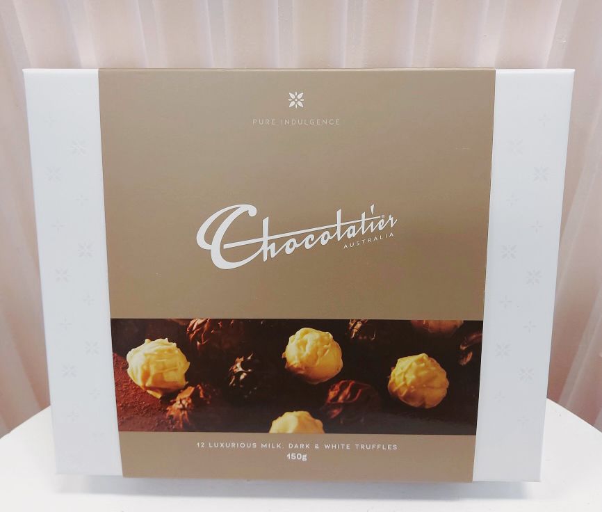 A box of Chocolatier brand chocolate truffles, with a picture of the truffles on a white background. The packaging is primarily white and brown with text and the Chocolatier logo. Bloom and Rocket Florist Taringa. Brisbane Flower Delivery.