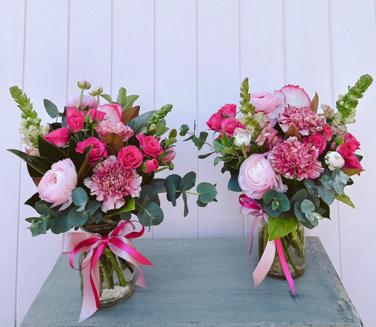 Two floral arrangements with vibrant pink flowers and green foliage, tied with pink.ribbons, displayed on a table. Dazzle me twice. Bloom and rocket florist. Brisbane same day flower delivery.