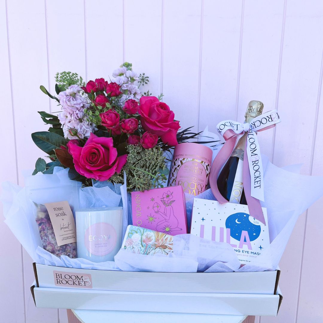 A gift basket containing an assortment of items including chocolates, a bottle of Chandon Sparkling Wine, eye masks, bath soap, body butter, and a candle, all arranged in a white box with a pink background. Brisbane gift delivery. Bloom and Rocket Florist.