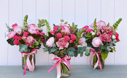 Three floral arrangements with pink and green flowers, tied with ribbons, placed in clear vases.